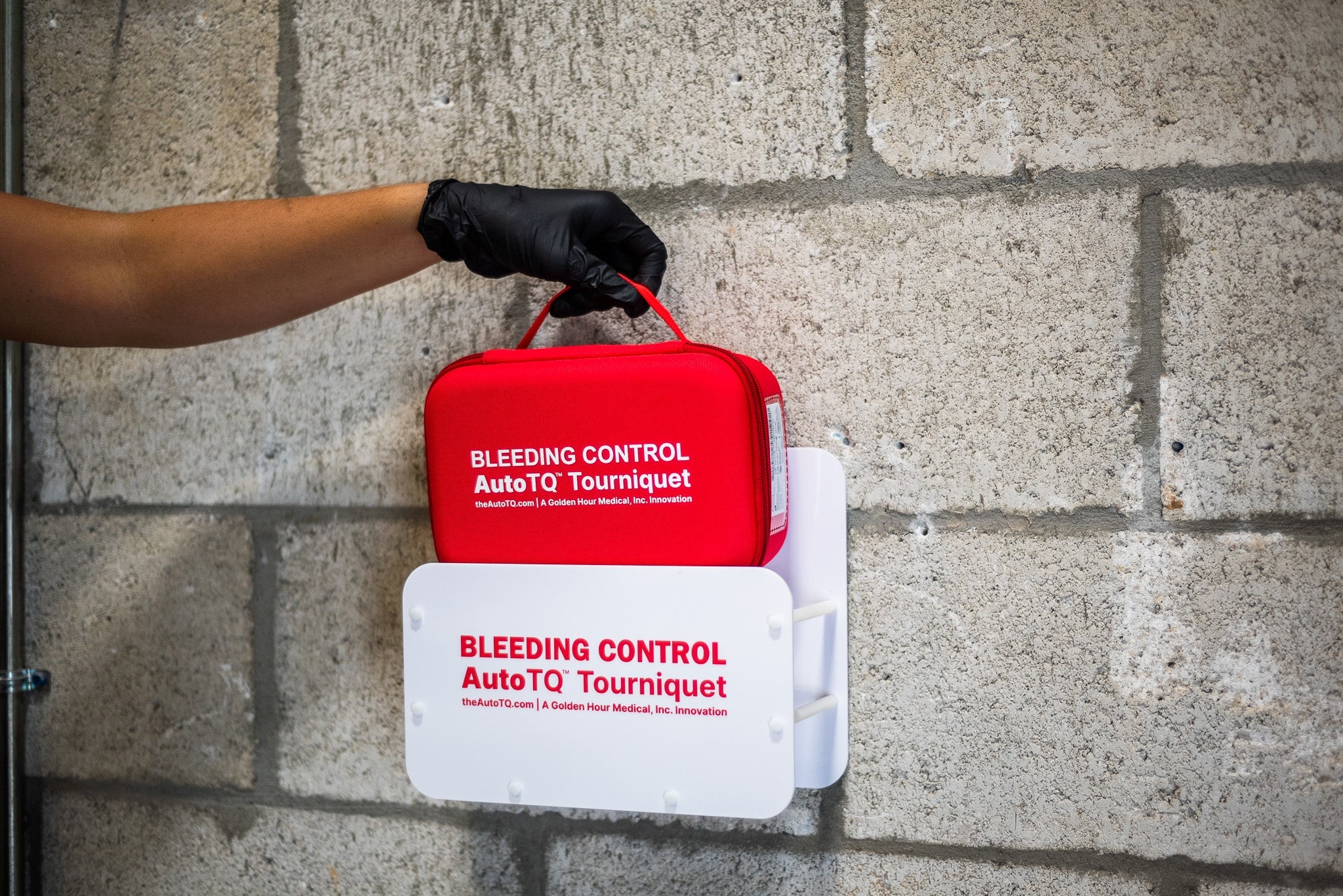 Red AutoTQ kit held by a gloved hand against a concrete wall.