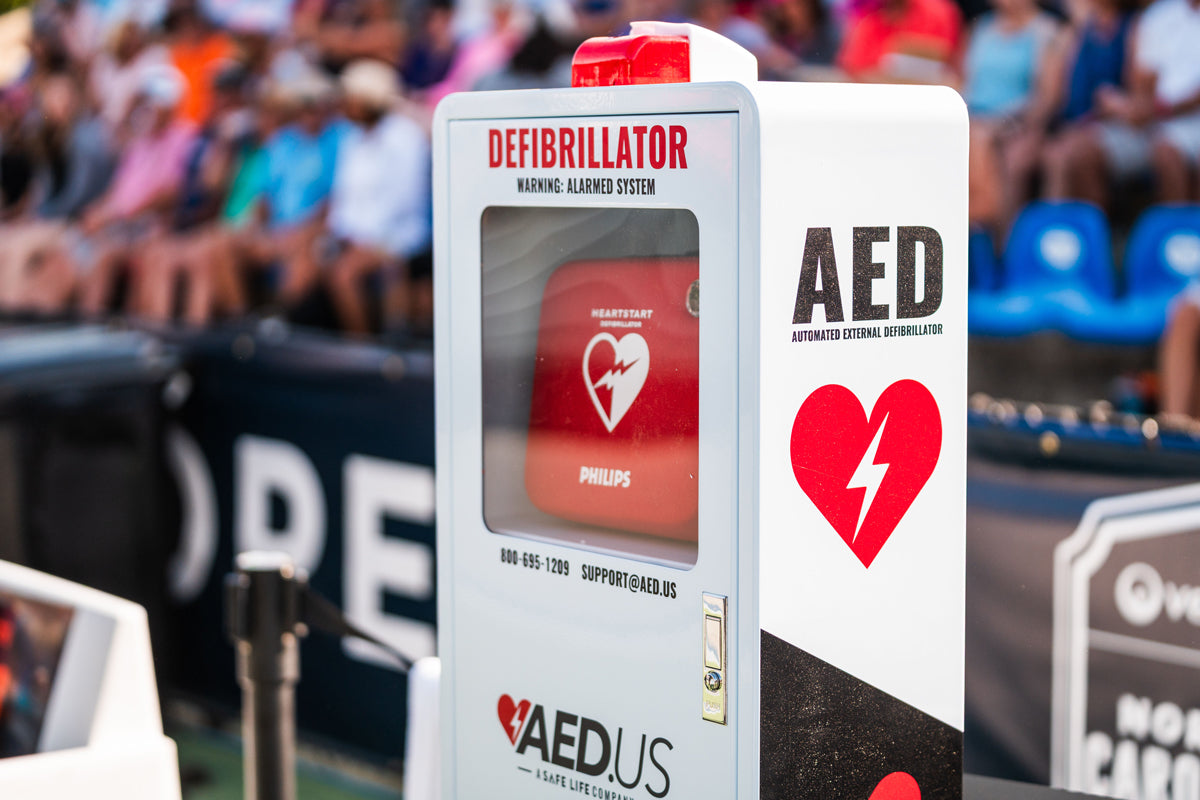 White AED cabinet sitting courtside at a PPA pickleball event. Inside is a red case with a Philips Onsite AED.