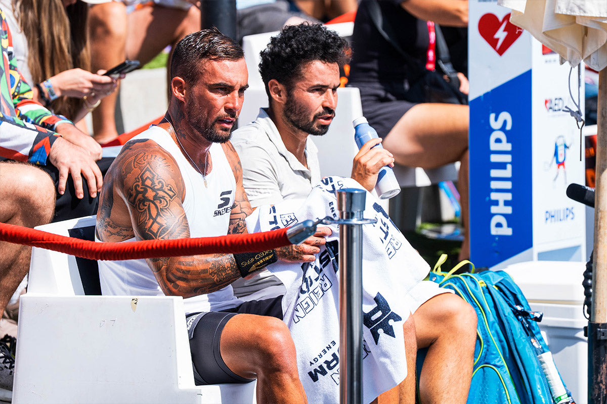 Two professional pickleball players sitting on a bench at the PPA Oragne County Cup with a Philips AED cabinet in the background. Tyson McGuffin in a white tank top holds a white towel, Noe Khlif sits beside Tyson about to drink from his water bottle.