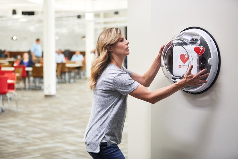 Woman using a wall-mounted device with a heart icon in an indoor setting.