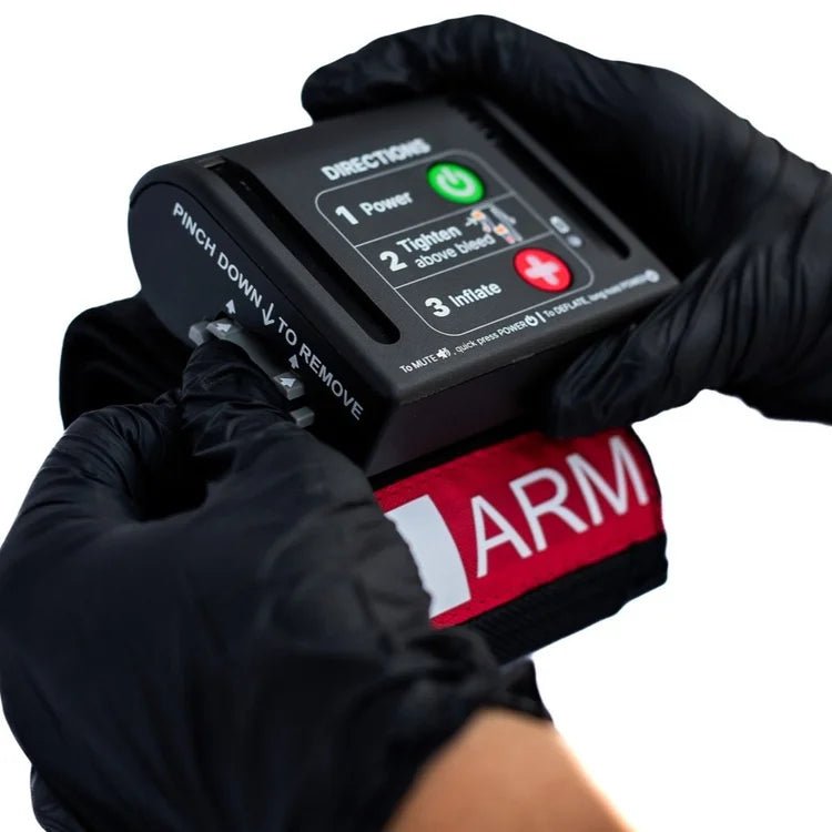 Person wearing black gloves holding a AutoTQ (automated tourniquet) device with 'ARM' branding on a white background